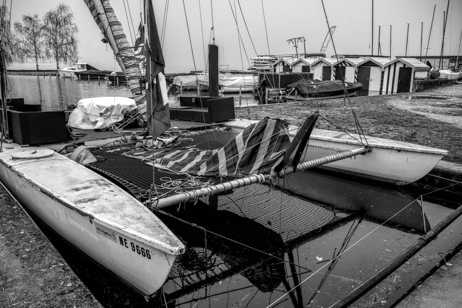 A black and white image of a catamaran docked at a marina with nearby storage sheds.